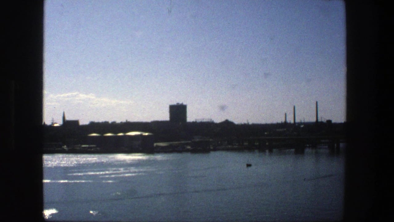 Stockholm waterfront in 1975 with reflective water, low industrial structures, smokestacks, and a prominent tower in silhouette.