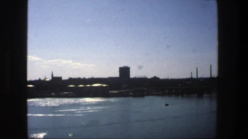 1975 Stockholm harbor view with sunlight on the water and a distant industrial skyline partly framed by dark window edges.