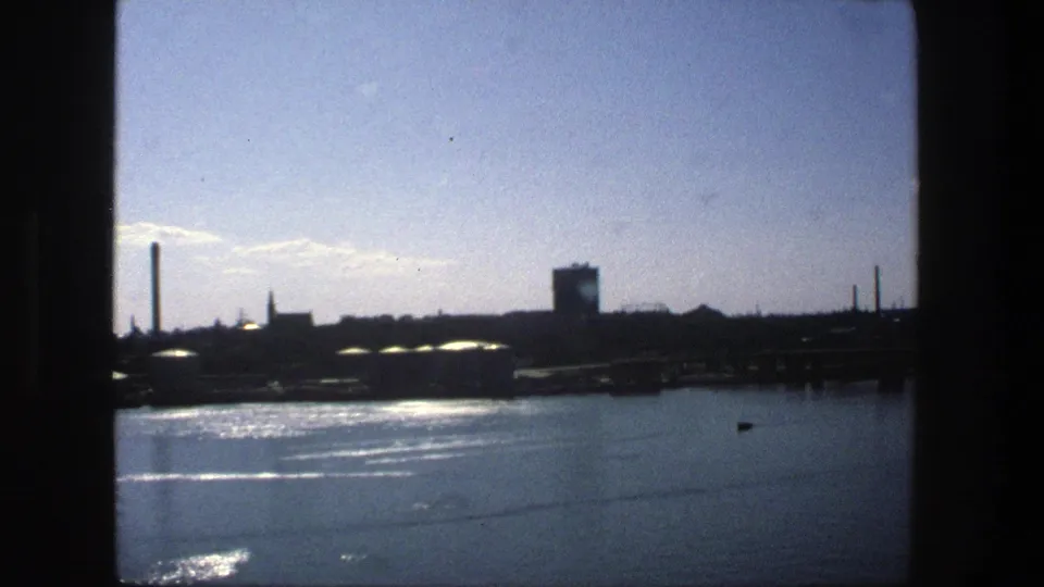 Late clip frame of the 1975 Stockholm waterfront, showing sunlit water, a tall chimney, a blocky tower, and low industrial buildings.