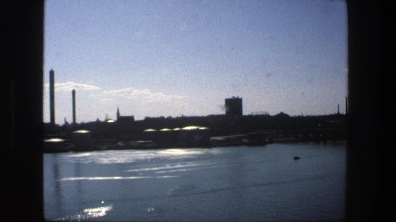 1975 Stockholm 8mm frame showing calm water, sun glare, and distant smokestacks silhouetted against a bright sky.