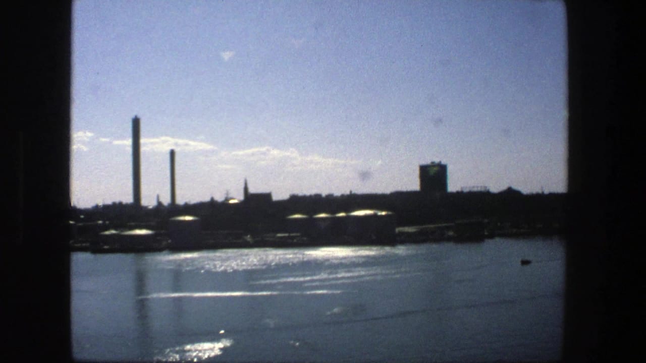 1975 Stockholm waterfront view with shimmering water, tall industrial stacks, storage tanks, and a bright blue sky.