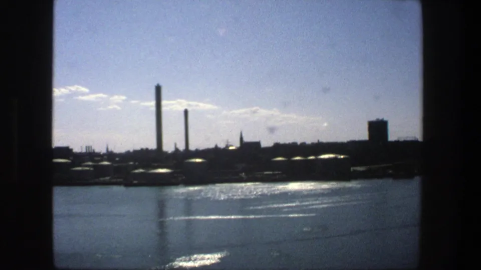 1975 Stockholm home-movie frame showing smokestacks, low waterfront structures, and sparkling harbor water.