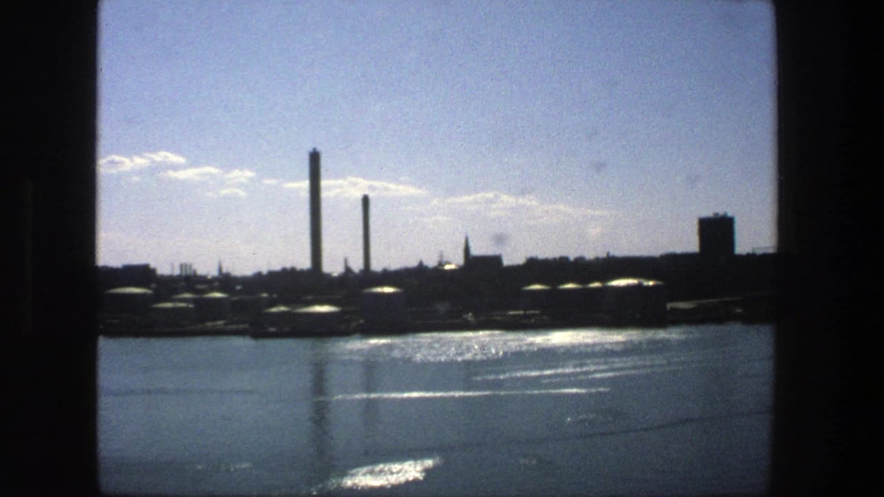1975 Stockholm waterfront view with reflective water, dark industrial buildings, tall smokestacks, and scattered clouds on 8mm film.