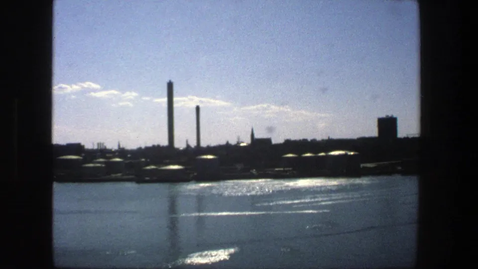 1975 Stockholm waterfront view with reflective water, dark industrial buildings, tall smokestacks, and scattered clouds on 8mm film.
