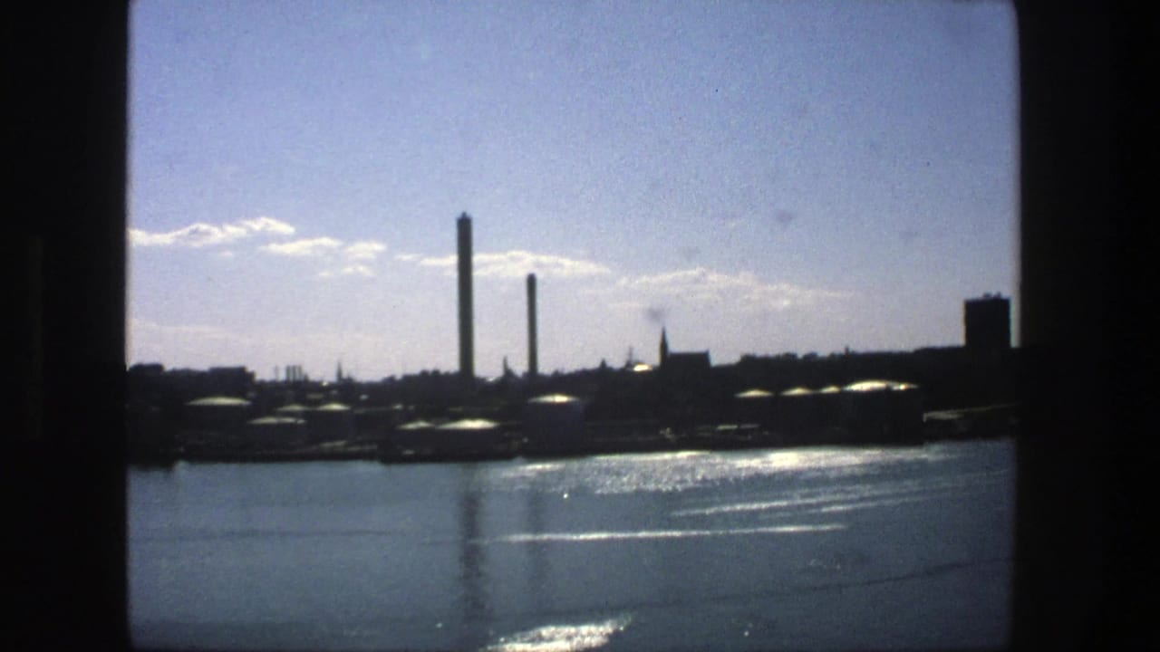 Industrial waterfront skyline across calm water, framed by dark film edges.