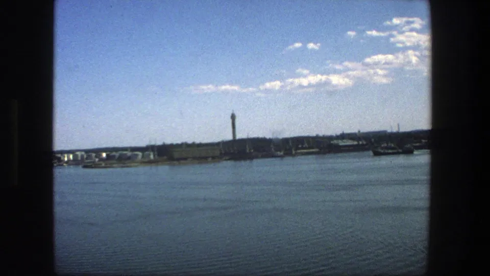 Stockholm waterfront in 1975 with calm blue water, distant industrial buildings, a tower, and bright cloud-dotted sky.