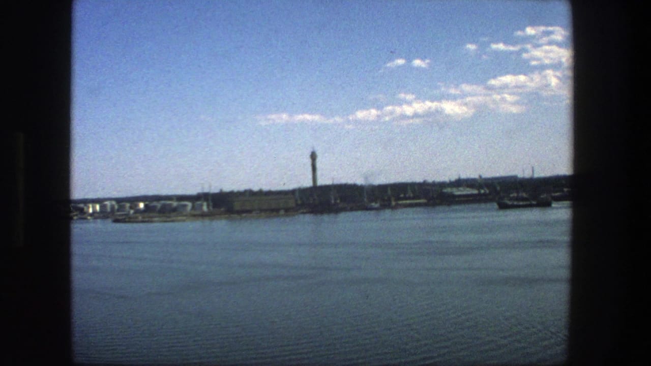 1975 Stockholm waterfront view on 8mm film, with calm water, distant buildings, a tall tower, and dark framing at the edges.