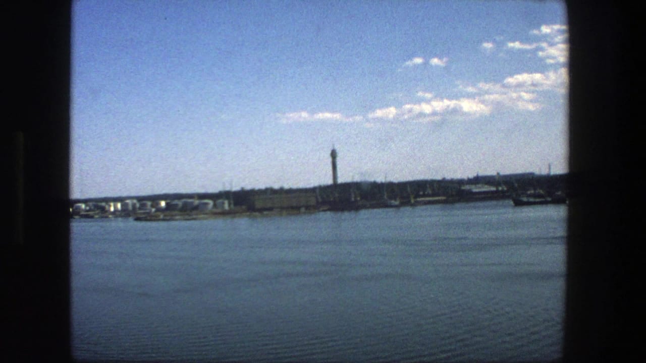 Stockholm waterfront in 1975, showing a broad expanse of water, a distant urban edge, and a prominent tower in soft 8mm color.