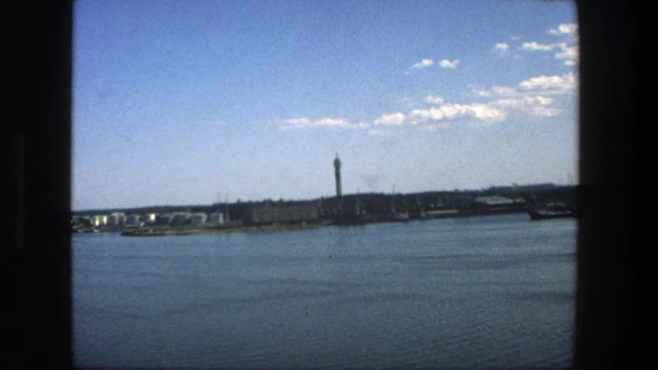 1975 8mm view of Stockholm across the water, with a distant tower, low buildings, rippled water, and dark window-like framing.