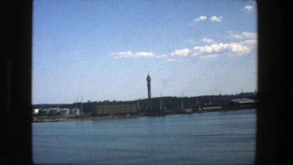 Stockholm waterfront view from 1975, with blue water in the foreground, a distant tower, low shoreline buildings, and vintage 8mm softness.