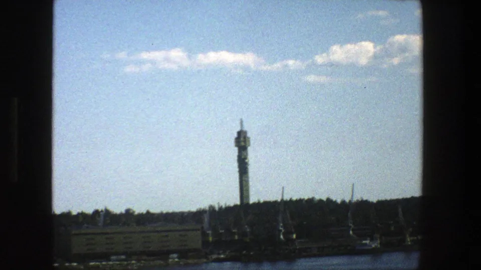 A tighter 1975 Stockholm home-movie frame centers the tower above the waterfront and wooded shore.
