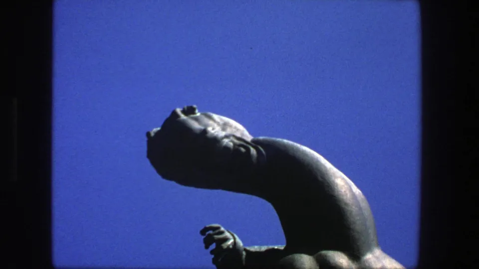 1975 Stockholm 8mm frame with a dark sculptural figure, tilted head, curved neck, and small hand form against blue sky.