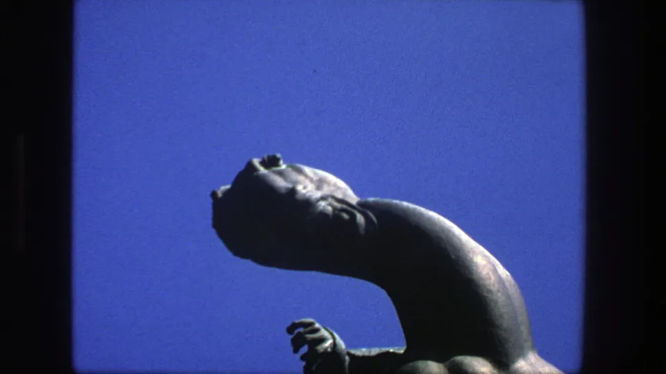 1975 Stockholm 8mm frame of a dark abstract statue, partly silhouetted, with head, neck, and small hand visible against blue sky.