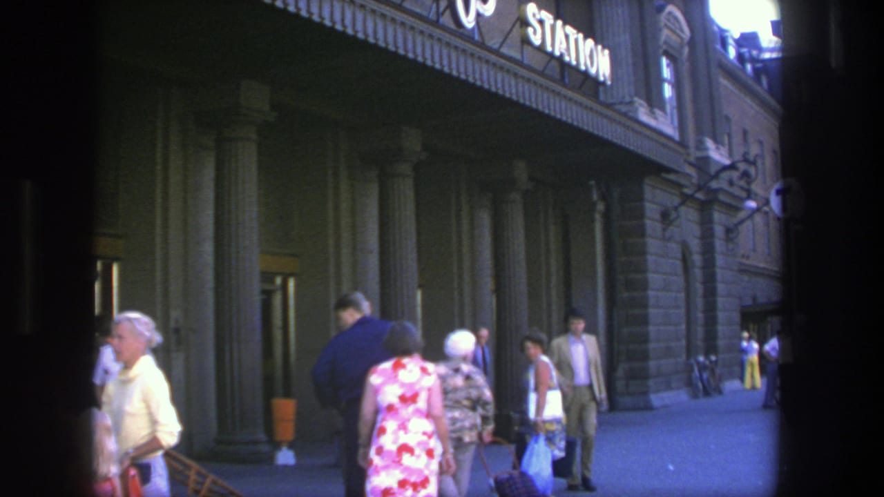 Stockholm station frontage in 1975 with adults walking near columns, luggage, and illuminated exterior signage.