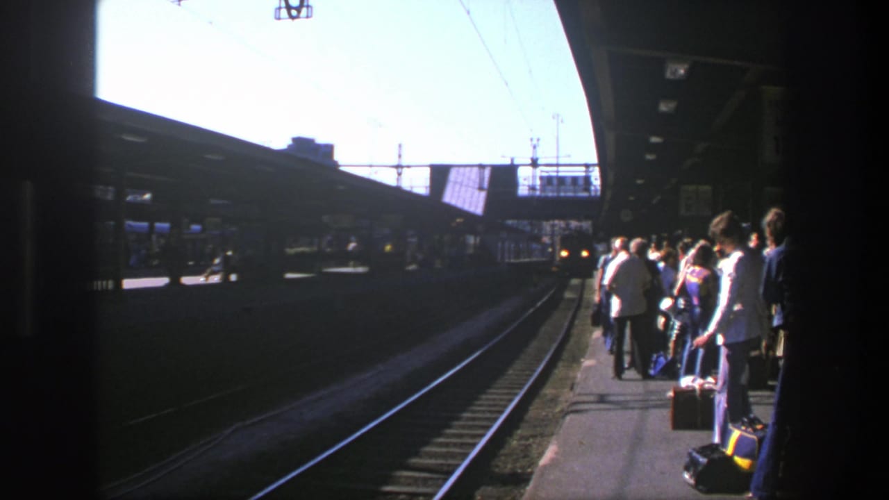1975 Stockholm train platform with passengers and luggage waiting beside tracks under overhead wires.