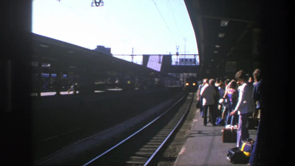 1975 Stockholm train platform with passengers and luggage waiting beside tracks under overhead wires.