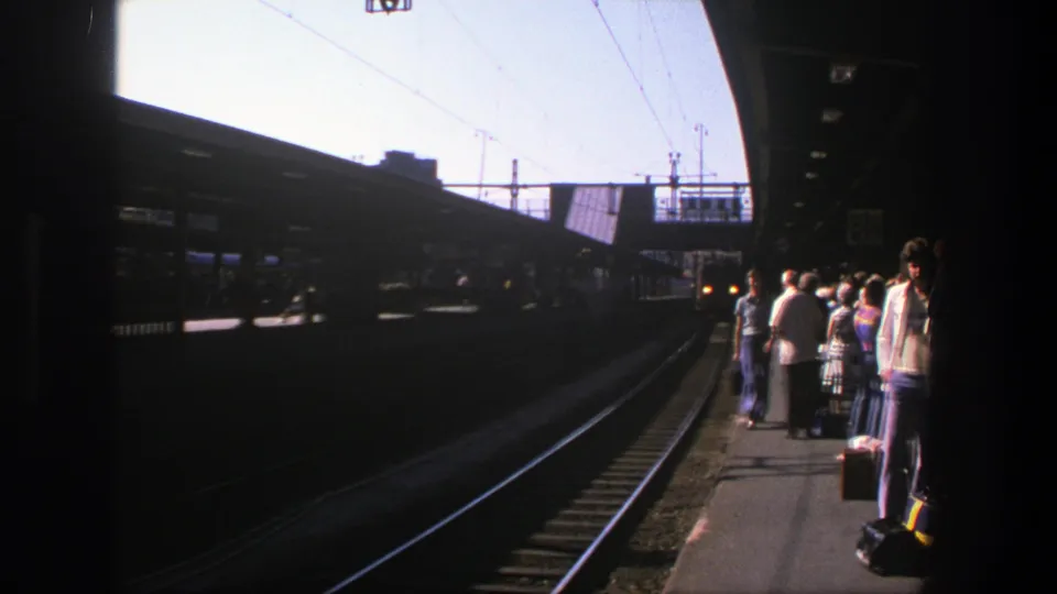 Sunlit 1975 Stockholm rail platform seen from track level, with passengers and luggage at the edge.