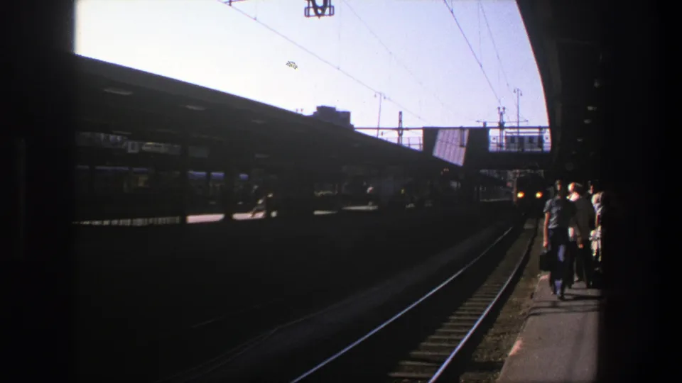Handheld 8mm view of a Stockholm platform in 1975, with travelers moving near tracks and luggage.
