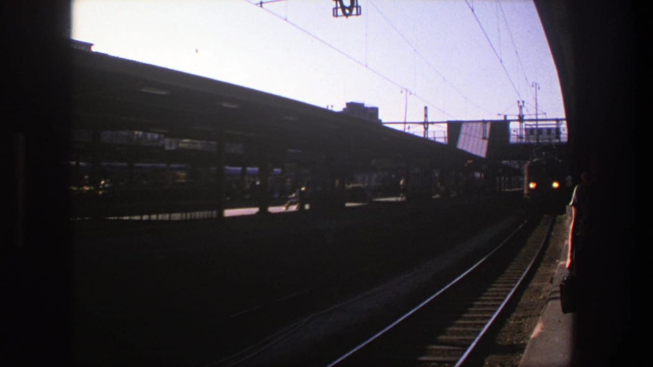 Stockholm rail platform in 1975 with dark foreground, overhead wires, station canopy, tracks, and silhouetted passengers in the distance.