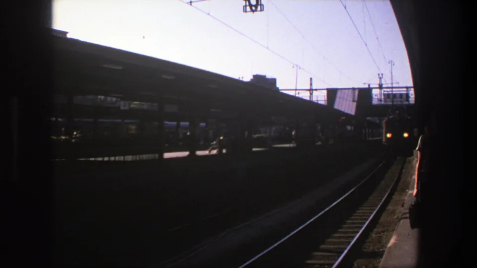 Stockholm rail platform in 1975 with dark foreground, overhead wires, station canopy, tracks, and silhouetted passengers in the distance.