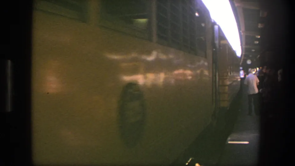 Side view of the yellow passenger train moving past the platform, with distant people waiting under the canopy.