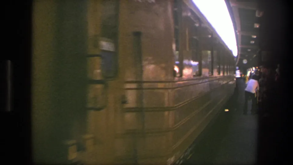 A yellow train car continues past a Stockholm platform while a person in a white shirt waits near the edge.