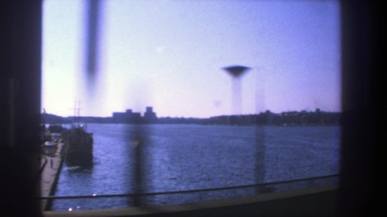Stockholm waterfront viewed through glass from a moving subway train, with harbor water, distant skyline, and a tall tower-like form beyond the window.