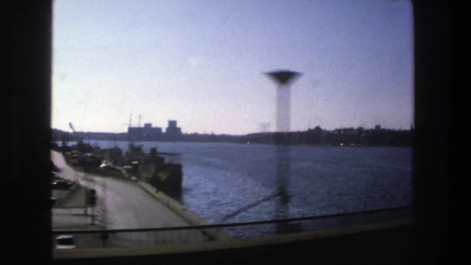 1975 Stockholm harbor view from a moving train window, showing water, a waterfront road, parked cars, distant buildings, and vertical glass artifacts.