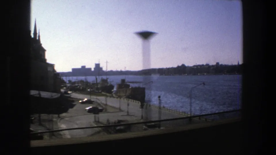 Stockholm waterfront from an elevated or moving train-window view, with road traffic, streetlights, harbor water, and a distant skyline in 1975 home-movie footage.
