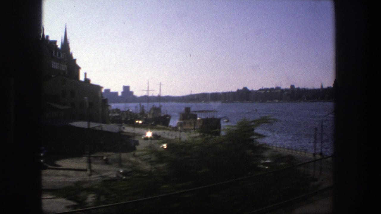 1975 Stockholm waterfront viewed from a moving train, with a pier, road traffic, docked boats, and dark window framing.