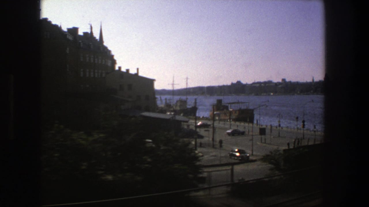 Stockholm waterfront in 1975 seen from transit, with a waterside road, small cars, pedestrians, buildings, and harbor water.
