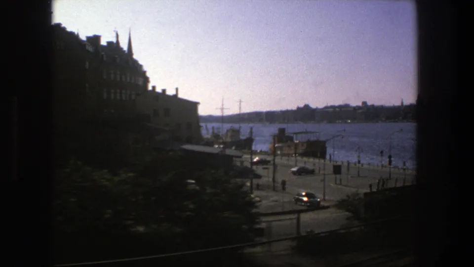 Stockholm waterfront in 1975 seen from transit, with a waterside road, small cars, pedestrians, buildings, and harbor water.