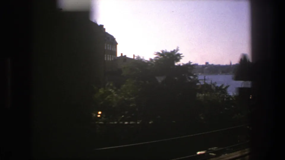 A 1975 Stockholm train-window view passes trees and waterside buildings, with harbor water and distant city forms visible.