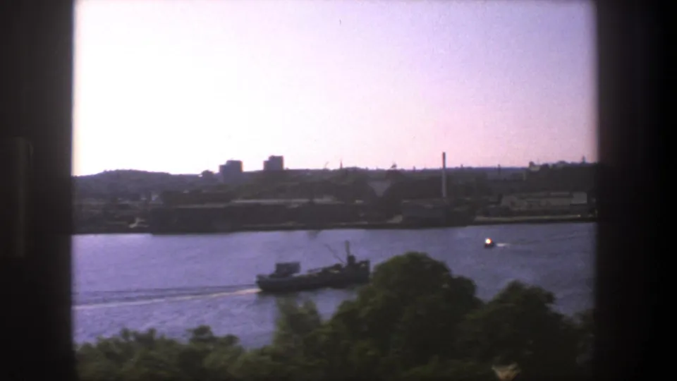 Stockholm waterfront seen from a moving train, with a small boat on the water, trees below, and an industrial-looking opposite shore.