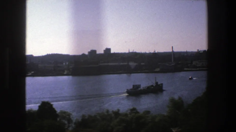1975 Stockholm harbor view from behind dark window edges, with a vessel crossing blue water below a silhouetted skyline.