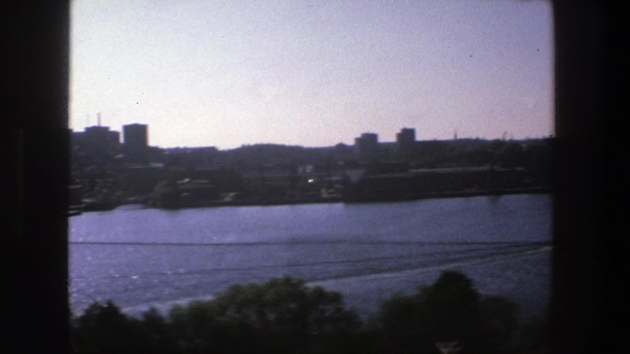 Window-framed 1975 Stockholm harbor view with blue water, trailing wakes, and distant buildings in silhouette.