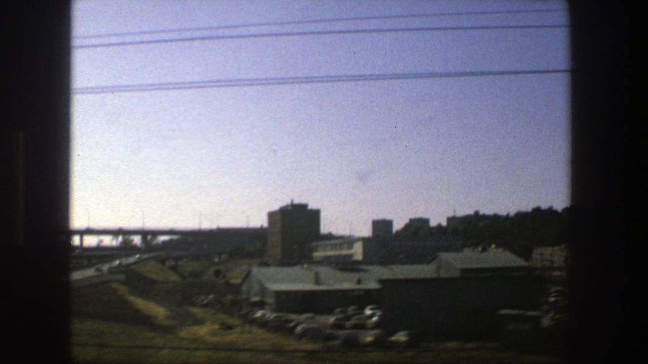 1975 Stockholm highway view from a moving vehicle, with an elevated bridge, low buildings, parked cars, and dark window borders.