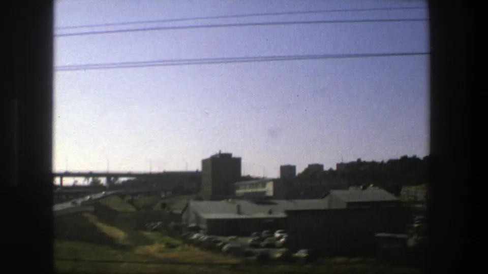 1975 Stockholm highway view from a moving vehicle, with an elevated bridge, low buildings, parked cars, and dark window borders.
