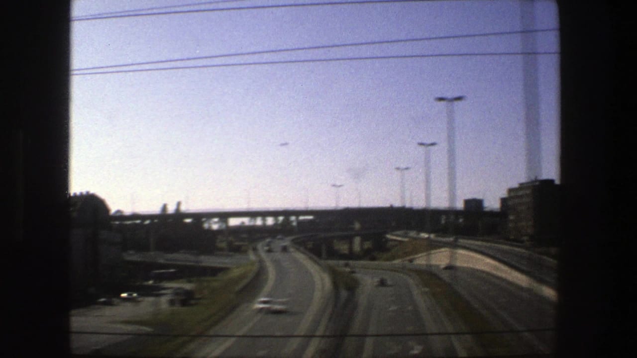 1975 Stockholm highway scene with divided lanes, a few cars, tall streetlights, an overpass, and a dark window edge in the foreground.