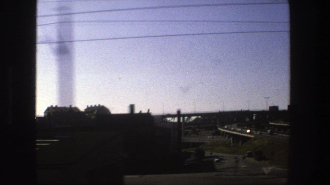 Late clip view of a Stockholm highway interchange in 1975, with overpasses, roadside structures, traffic, and dark vehicle-window framing.