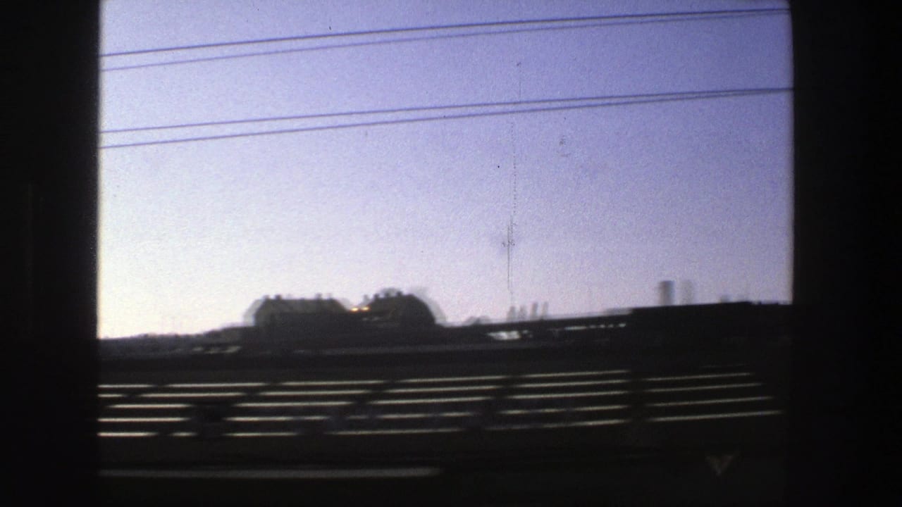 Dark vehicle window frame over a pale sky, power lines, roadside barriers, and distant buildings.