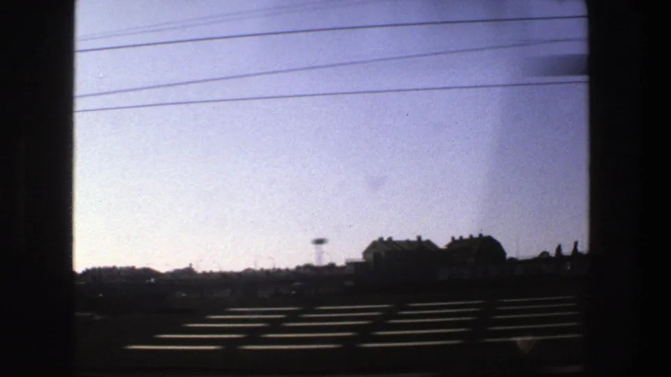 Stockholm-area roadside landscape seen from a moving vehicle in 1975, with power lines, low buildings, and dark foreground barriers.