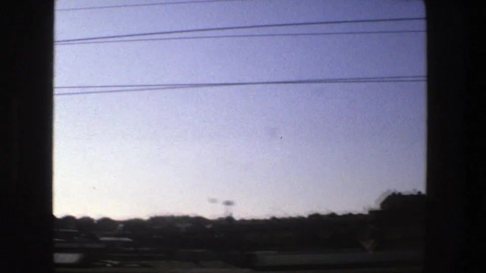 Passenger-window view of a Stockholm roadside in 1975, showing open sky, wires, dark trees, barriers, and small distant structures.