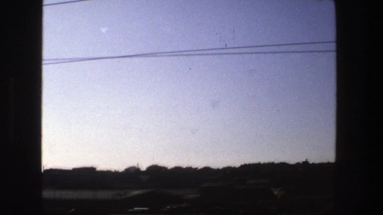 1975 Stockholm train-window view with dark frame edges, overhead cables, and a silhouetted urban skyline under a hazy sky.