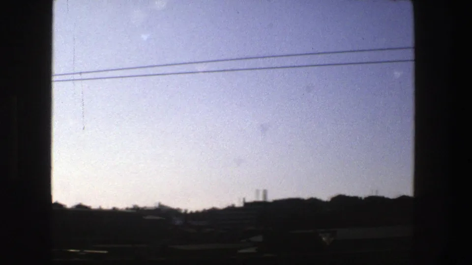 1975 Stockholm home-movie frame showing rail-side cables, dark window borders, and a distant skyline with rooftops and trees.