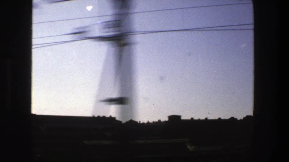 1975 Stockholm train-window view with a silhouetted skyline, overhead wires, and bright clear sky on small-gauge film.