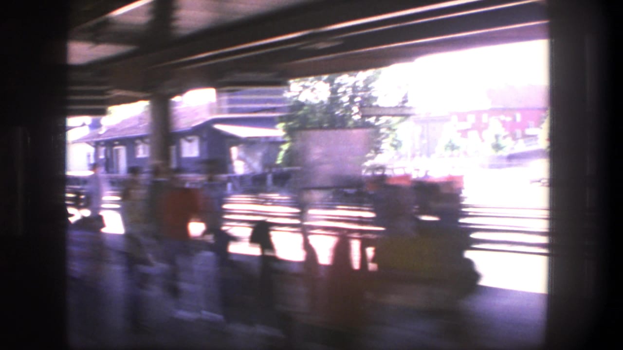 Blurred station platform seen from a moving train, with tracks, roof supports, and waiting figures.