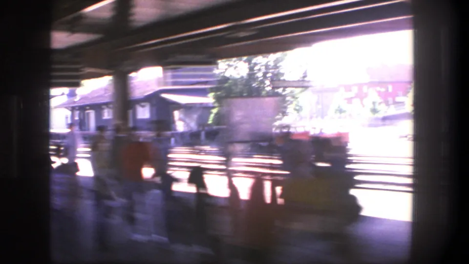 1975 Stockholm rail platform viewed through a moving train window, with motion blur, tracks, roof supports, and passengers waiting.