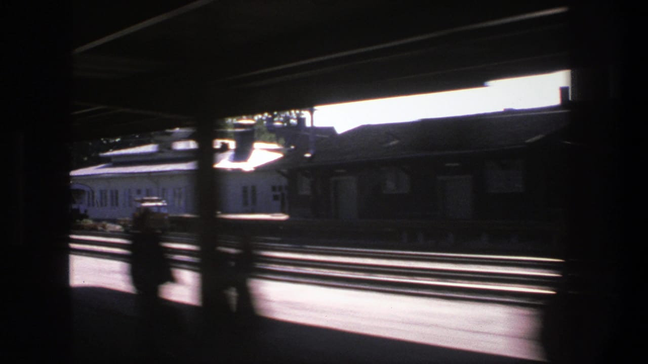 Motion-blurred 1975 Stockholm train view across multiple tracks, with silhouetted passengers and low station buildings beyond.