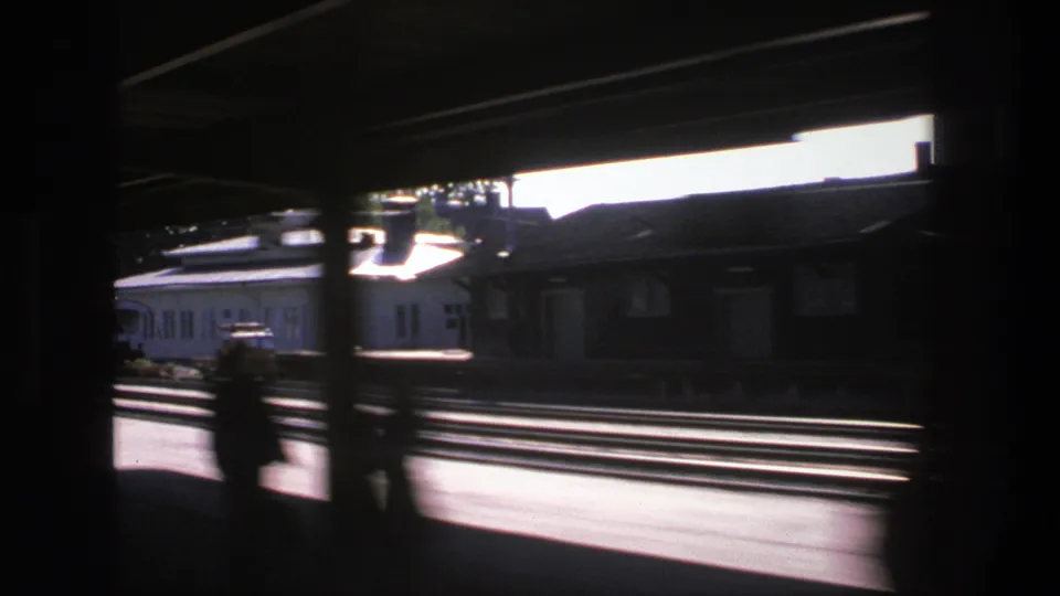 Motion-blurred 1975 Stockholm train view across multiple tracks, with silhouetted passengers and low station buildings beyond.